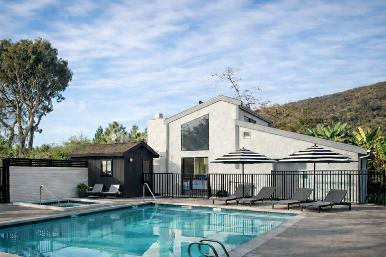 Swimming pool with lounge chairs, fenced area, white building, and mountain backdrop.