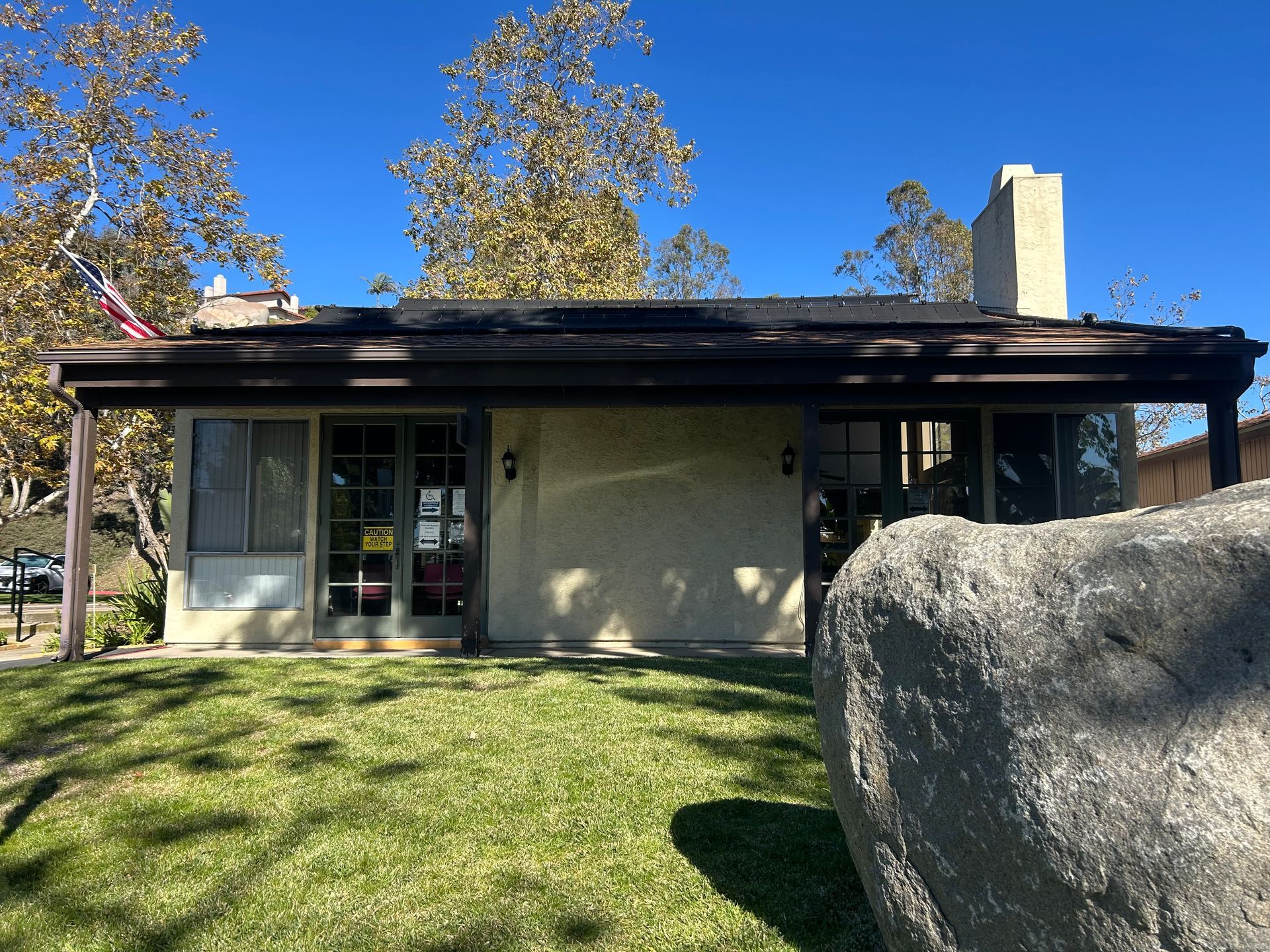 Small building with a brown roof and a large rock in front of it. Bright green grass and blue sky.
