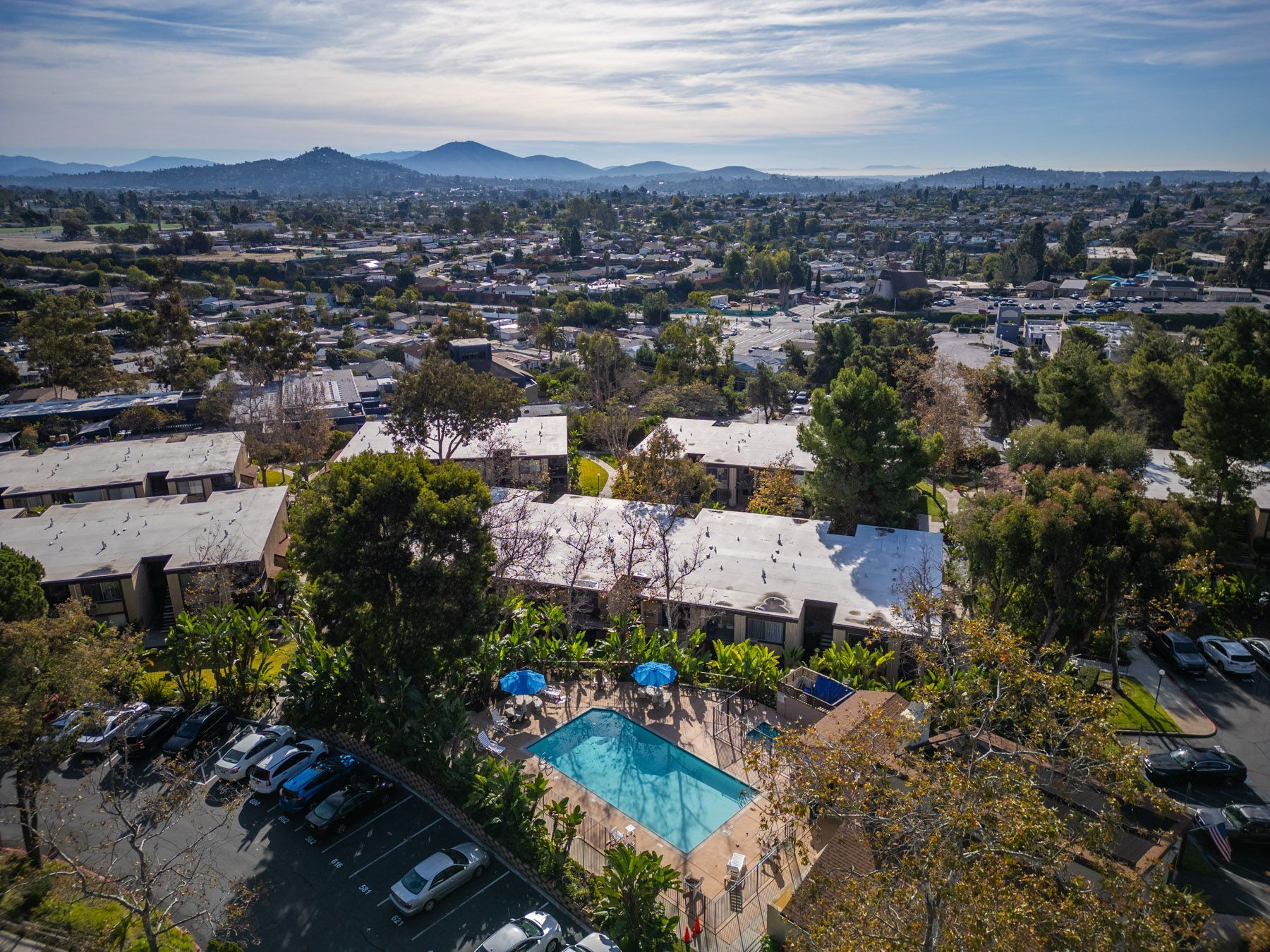 Aerial view of a complex at Cowles Landing with a pool, surrounded by buildings, trees, and a city with mountains in the distance.