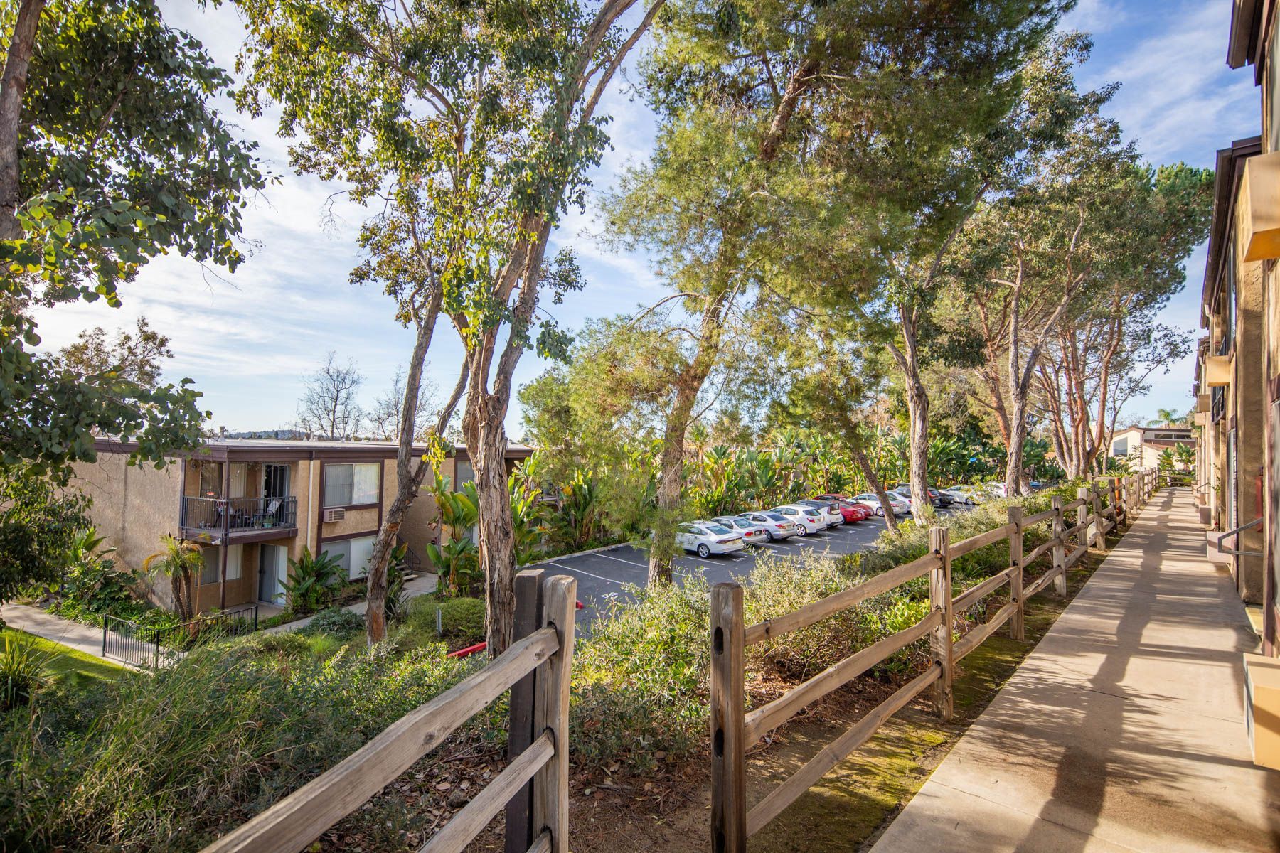A wooden fence borders a pathway beside a building, with trees and parked cars visible in the distance at Cowles Landing, offer apartments for rent in San Diego, CA.