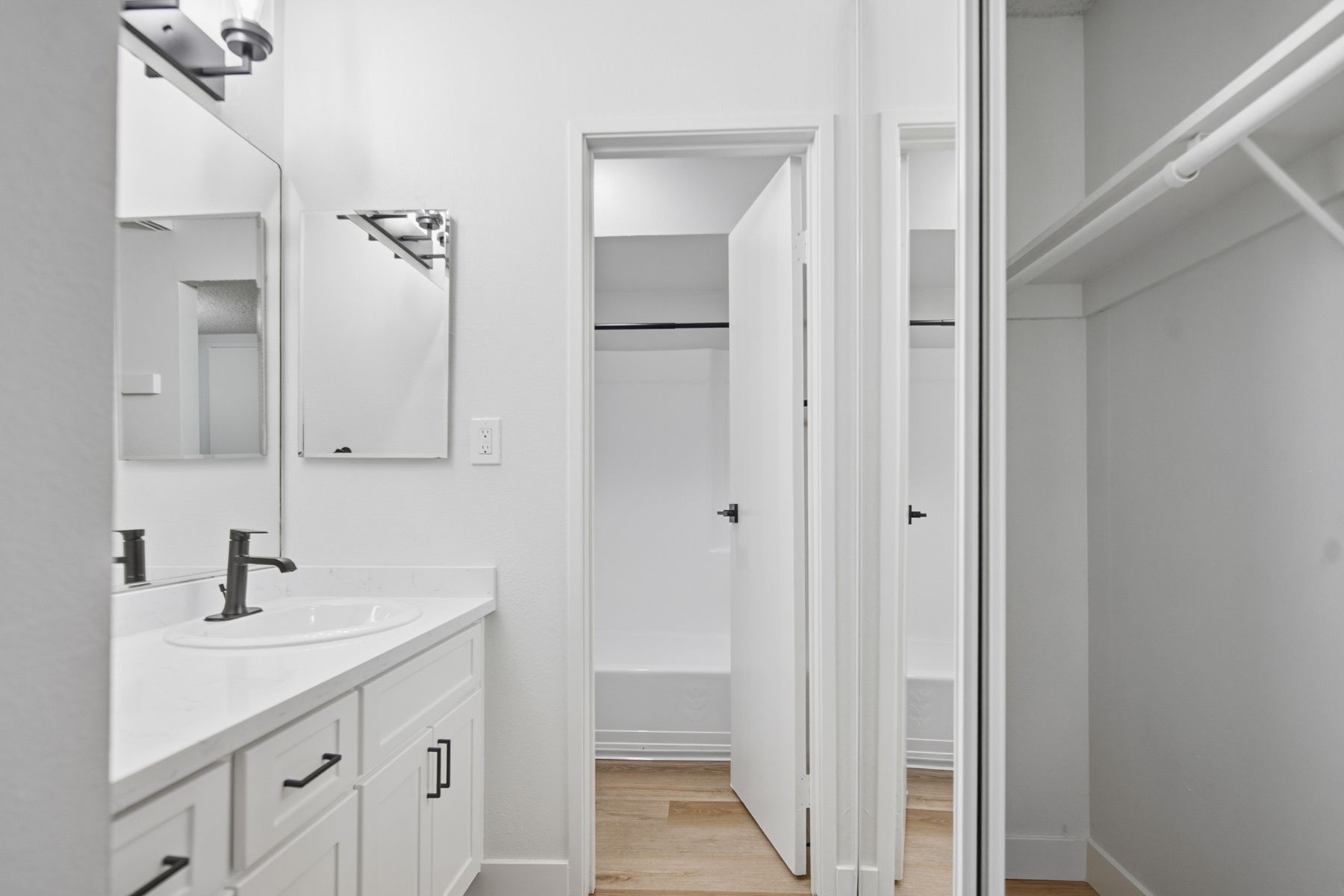 White bathroom with vanity, mirror, shower, and closet; light wood floor.