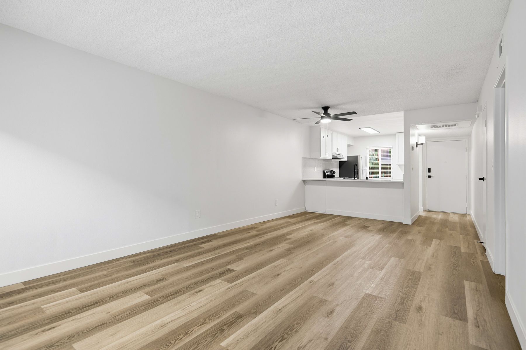 Empty living room with wood flooring, white walls, and a small kitchen visible in the background.