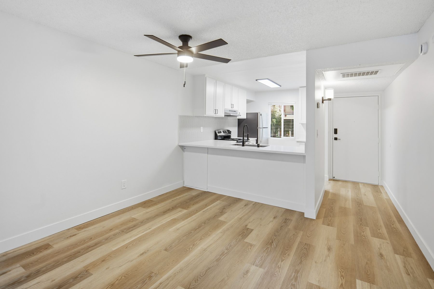 Empty white apartment with light wood floors and a partial kitchen.