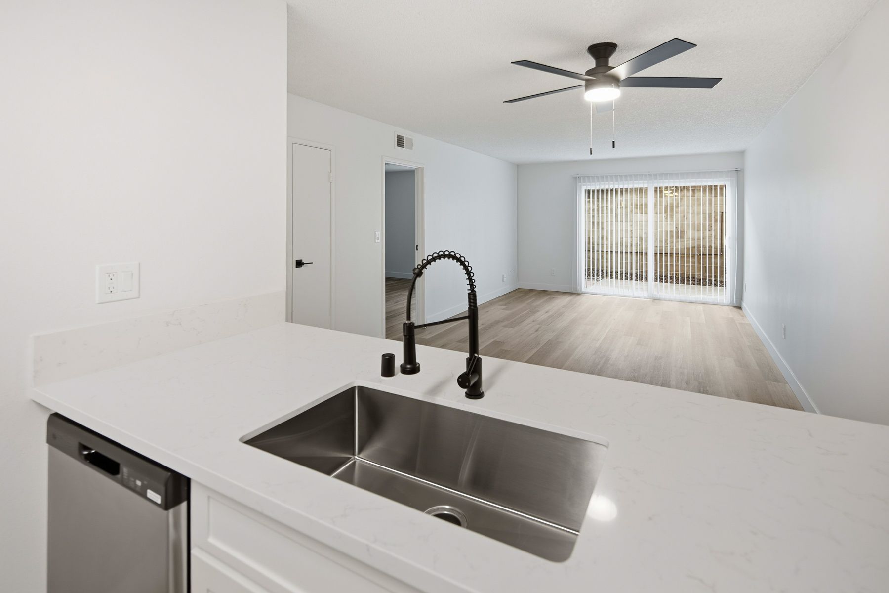 Kitchen with white countertop, black faucet and sink, leading to a living room with sliding doors and a ceiling fan.