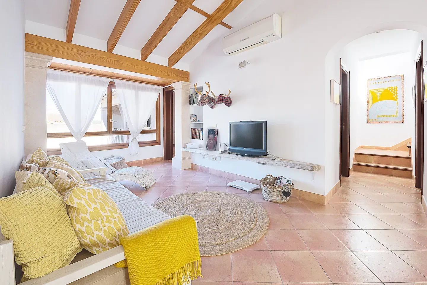 Sala de estar con paredes blancas, vigas de madera, un sofá con cojines amarillos y un televisor.