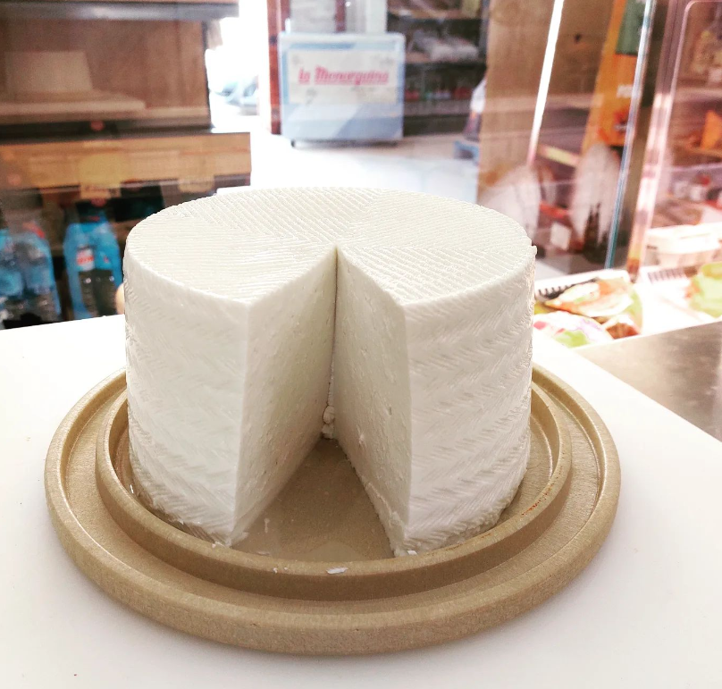 Gran rueda de queso blanco en una bandeja redonda de madera, con una cuña quitada. Interior de la tienda.