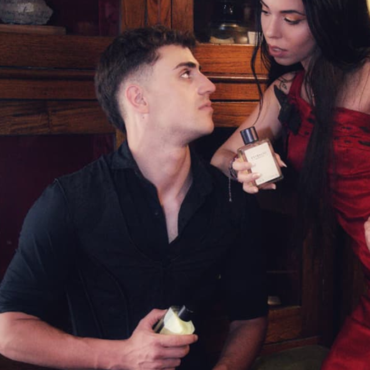 Un hombre mira a una mujer con perfume. Él sostiene un objeto blanco. Interior con iluminación cálida.