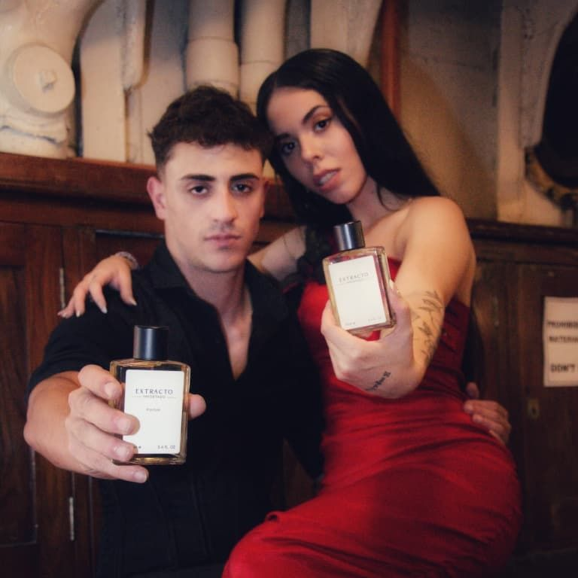 Un hombre y una mujer sostienen frascos de perfume. Están en un interior frente a un revestimiento de madera.