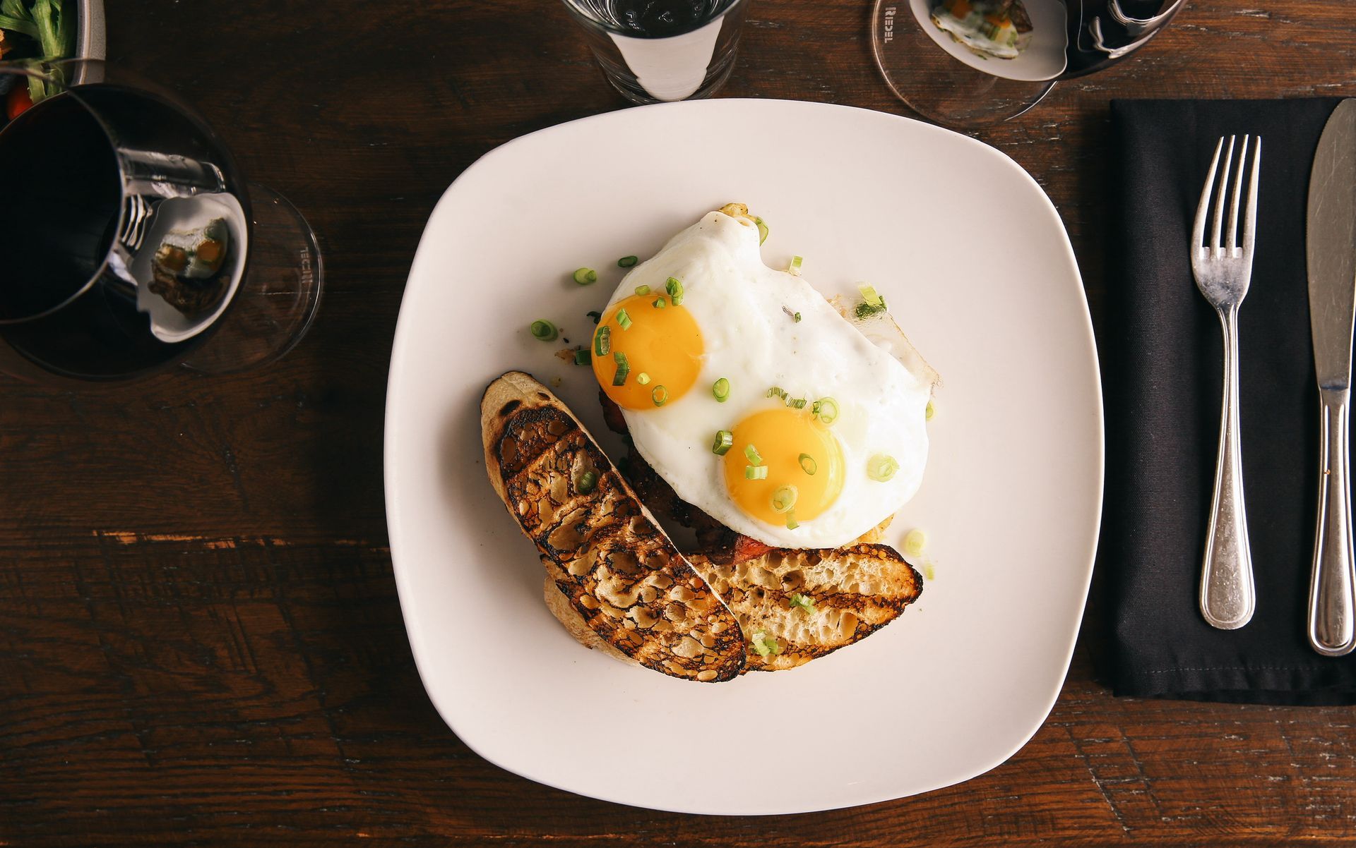 Overhead view of a plate with two sunny-side-up eggs and grilled bread, alongside silverware and a glass on a wooden table.