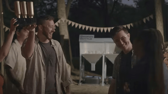 People at an outdoor party, toasting with drinks. Trees, bunting, and a keg are visible.