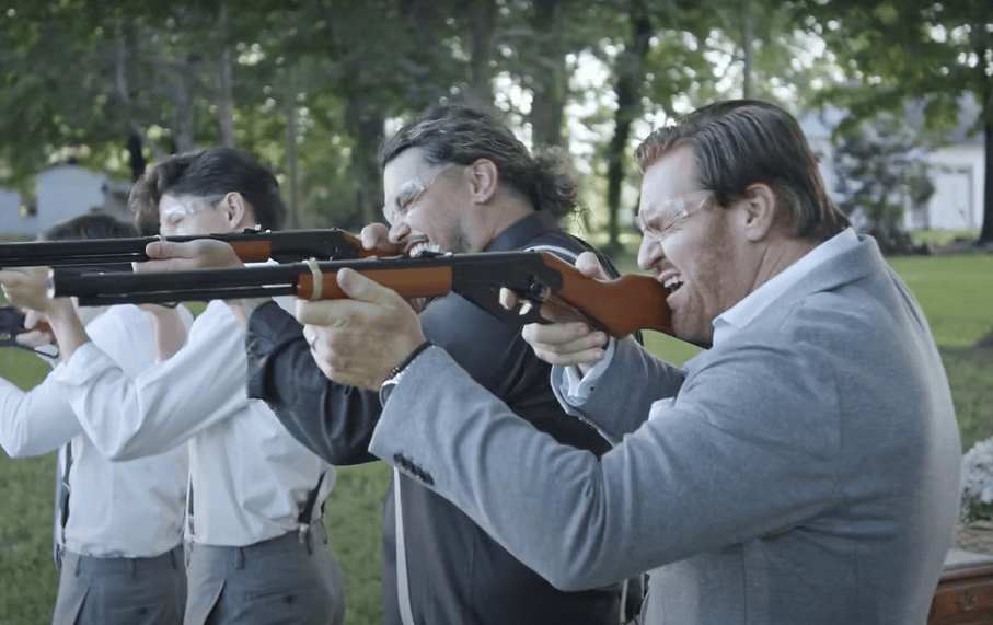Men in a row, aiming rifles. Setting is outdoors, with green grass and trees. One man is yelling.