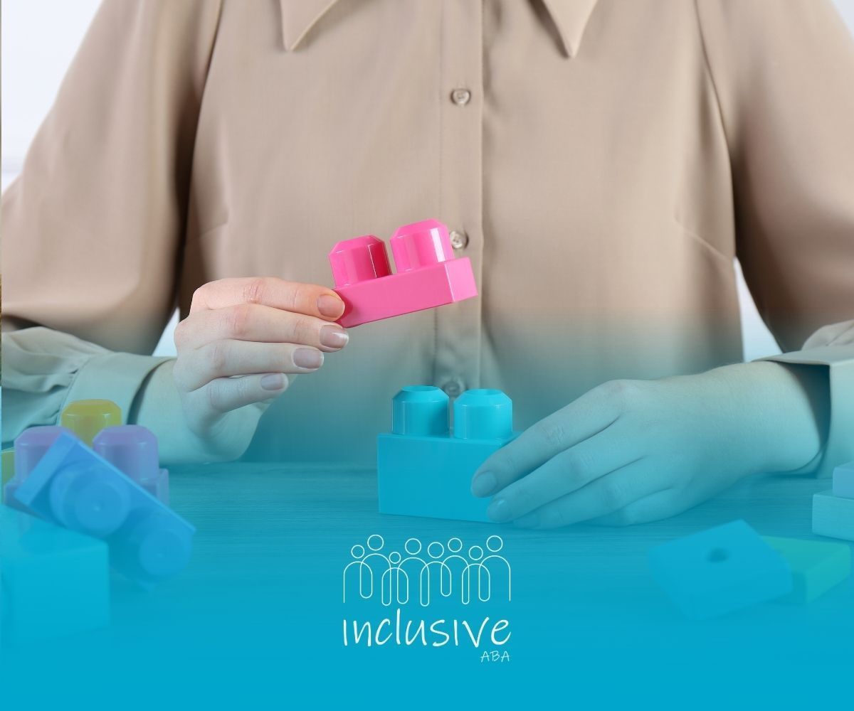 A person holds a pink toy block above a blue one on a wooden table, with other colorful building blocks scattered nearby.