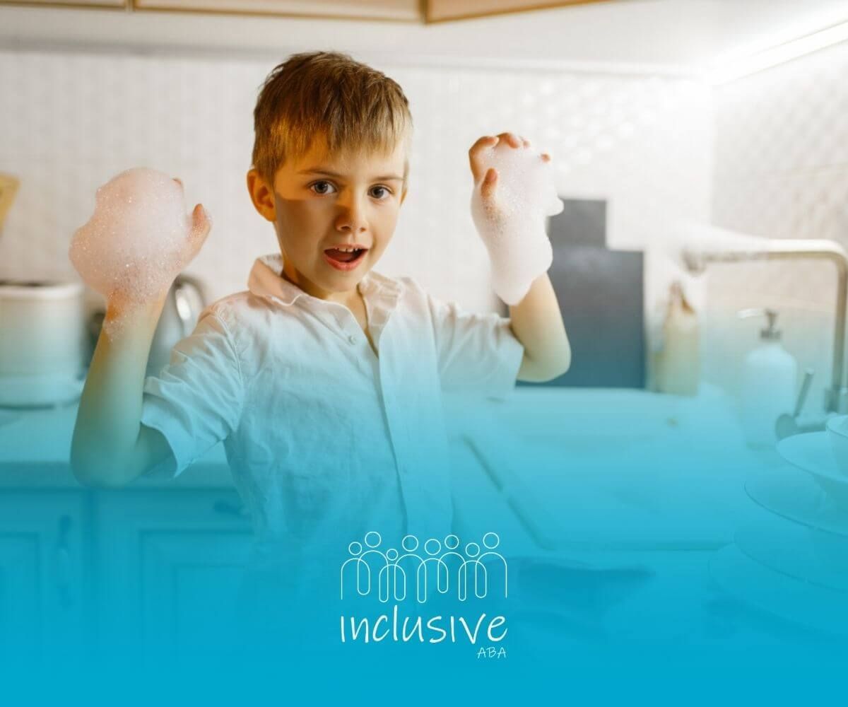 A boy with soapy hands, smiling while standing in a kitchen sink area.