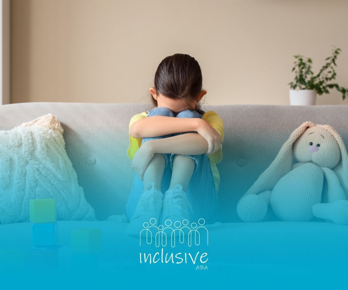 Girl sitting on a couch, knees to chest, head down. A toy rabbit sits beside her.