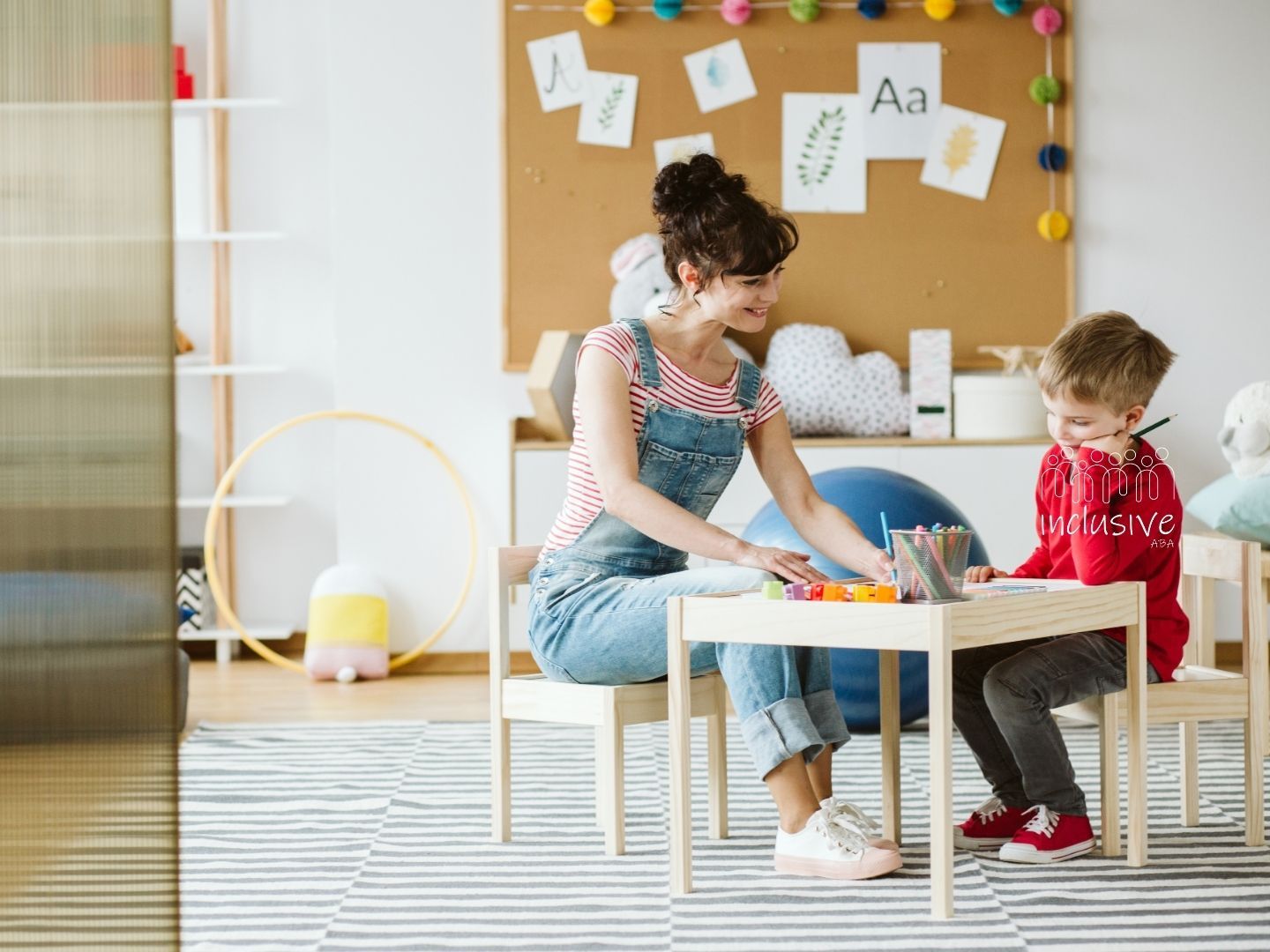 A teacher and student sit at a table in a classroom, drawing together on paper.