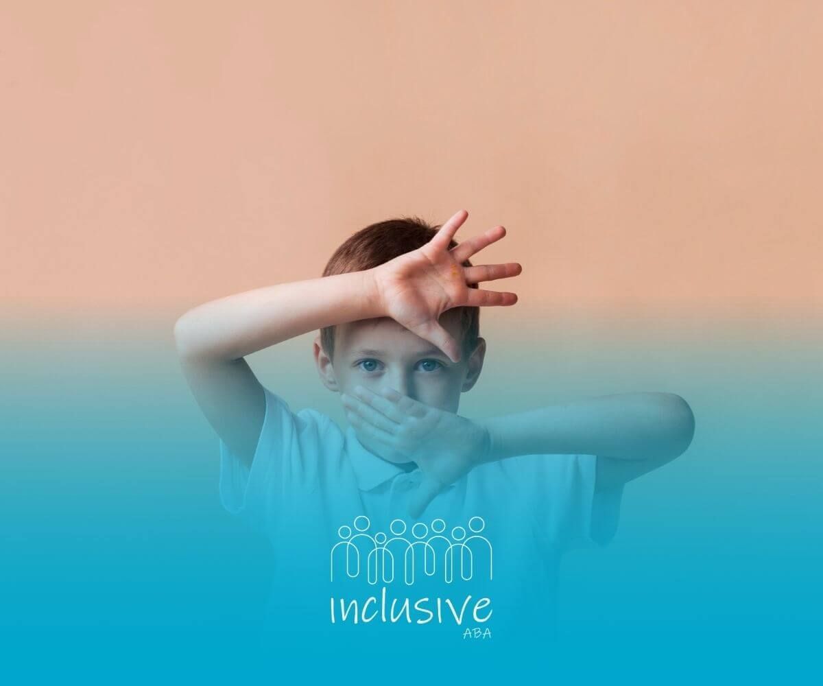 A child with autism covering their face with hands in front of a neutral-colored background.