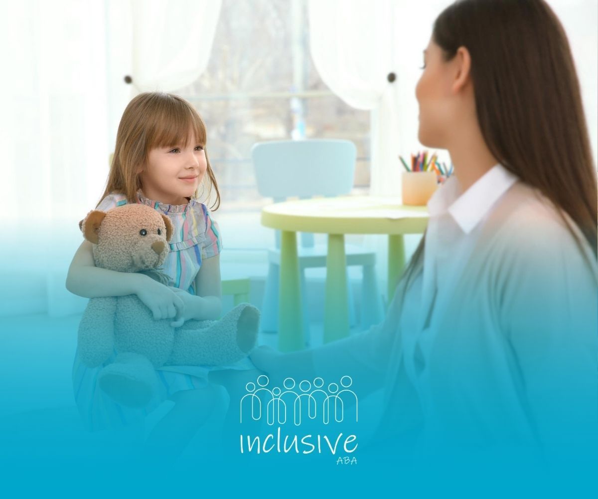 A BCBA is sitting next to a little girl holding a teddy bear.