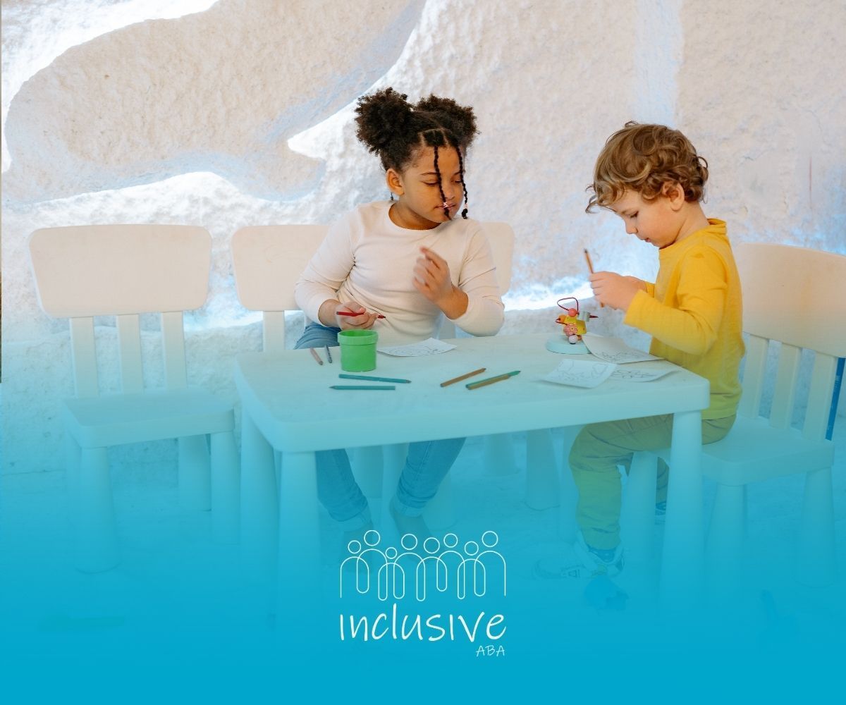 Two children sit at a table in a white, textured salt room, engaged in an activity with colorful art supplies.