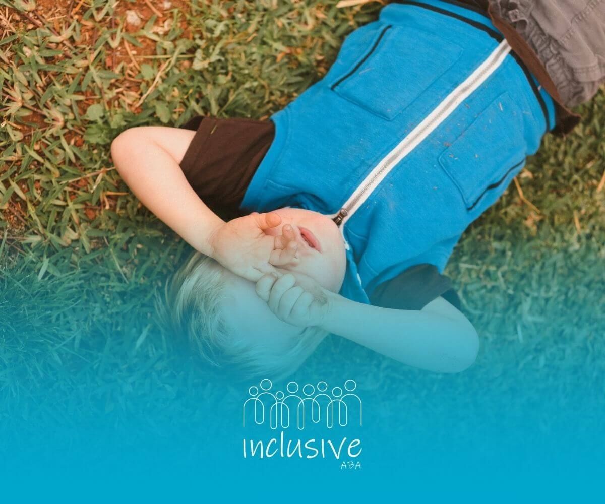 A boy with autism lying on the grass with his hands covering his face, looking up.