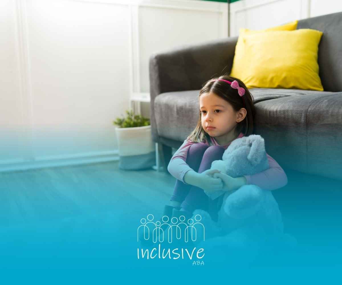 Young girl with sad expression hugging a stuffed animal, sitting on the floor near a sofa.