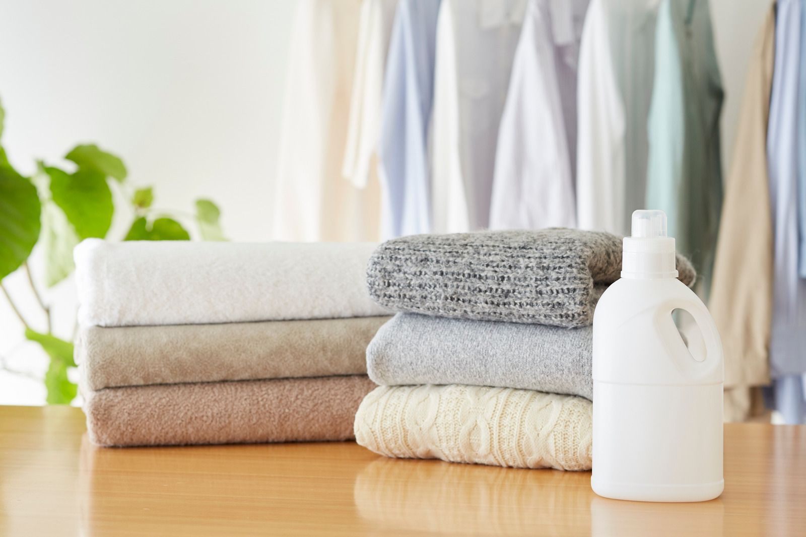 Stacks of folded towels and knit sweaters on a wooden table next to a white laundry detergent bottle.