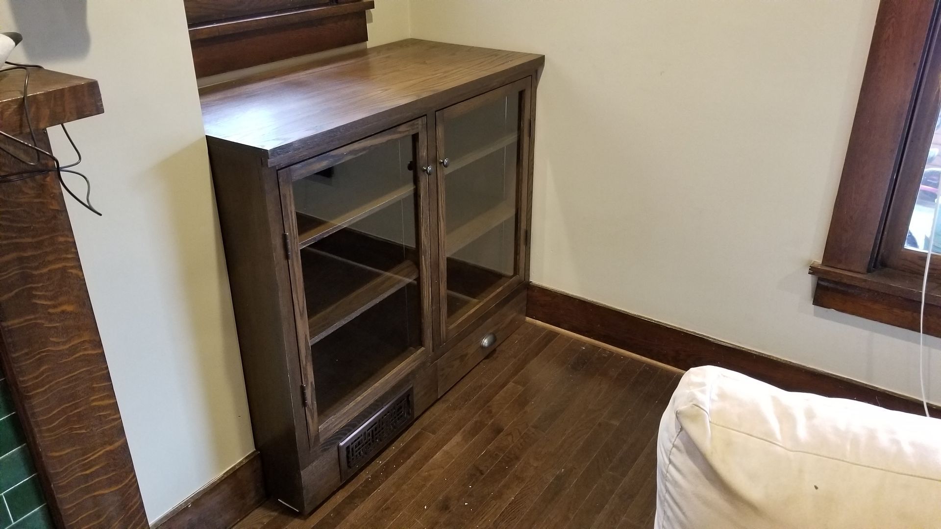 A wooden cabinet with glass doors is in a living room next to a window.