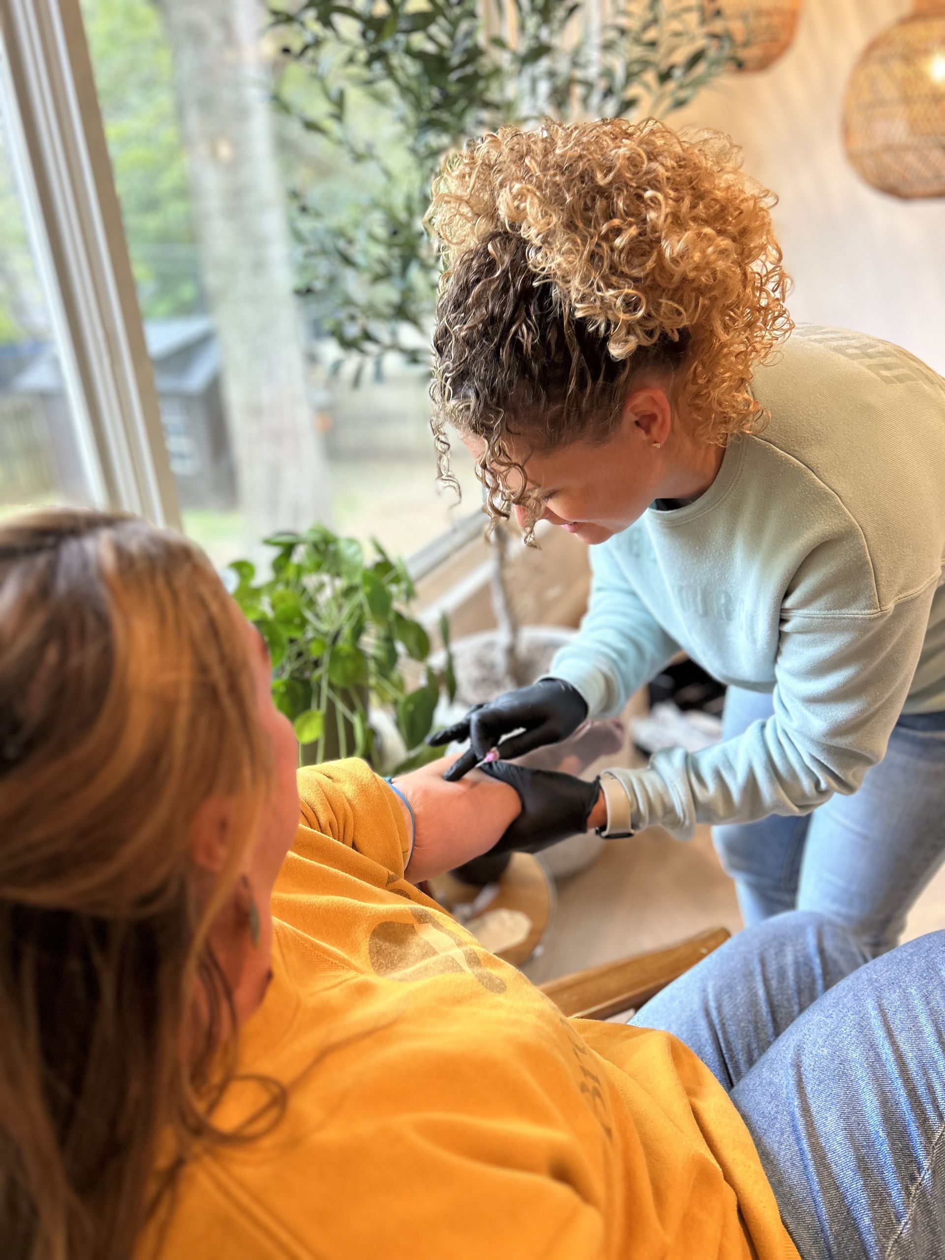 Woman applying something to another woman's arm, both near a window, natural light.