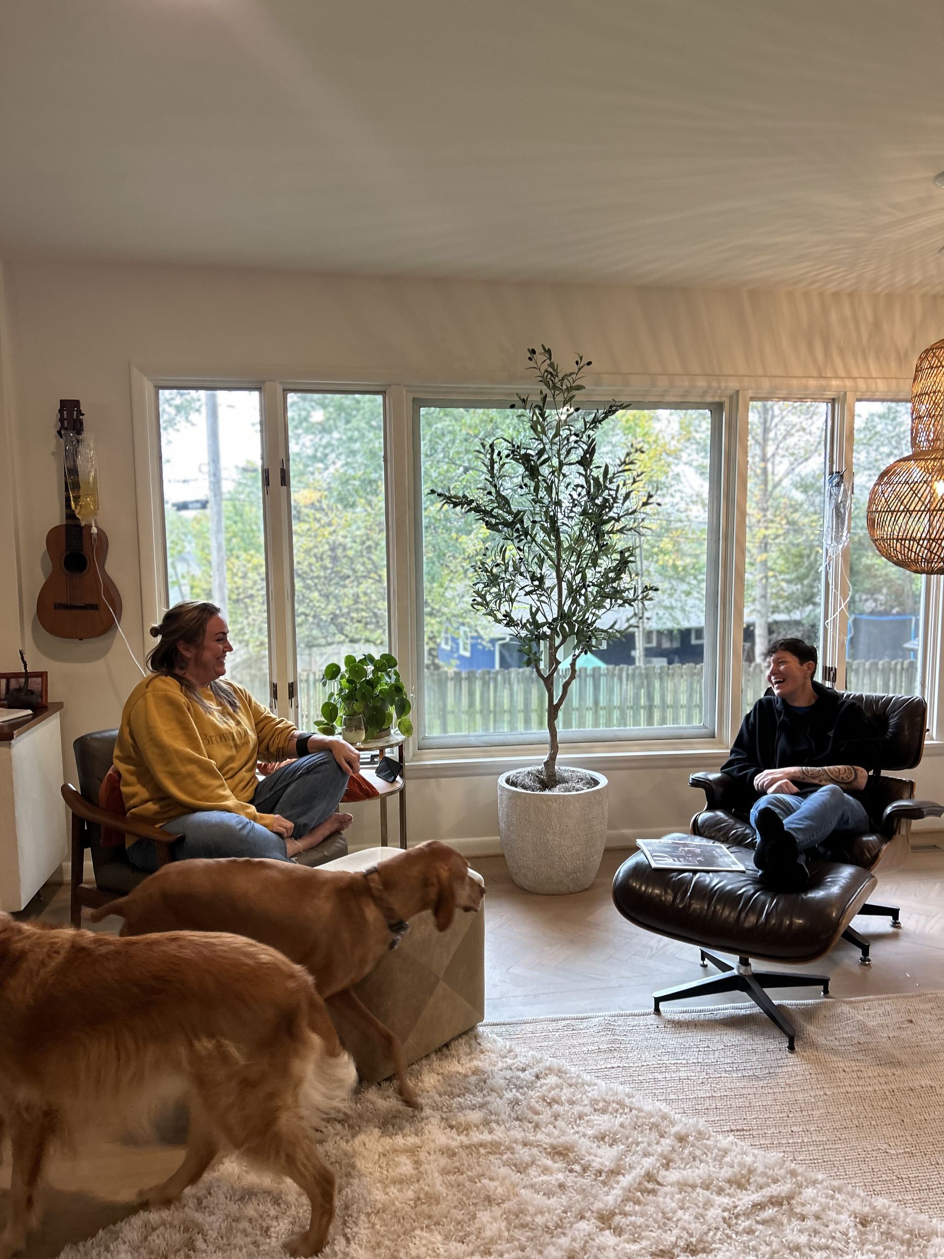 Two people seated, conversing in a living room with dogs, a tree, and a large window overlooking trees.