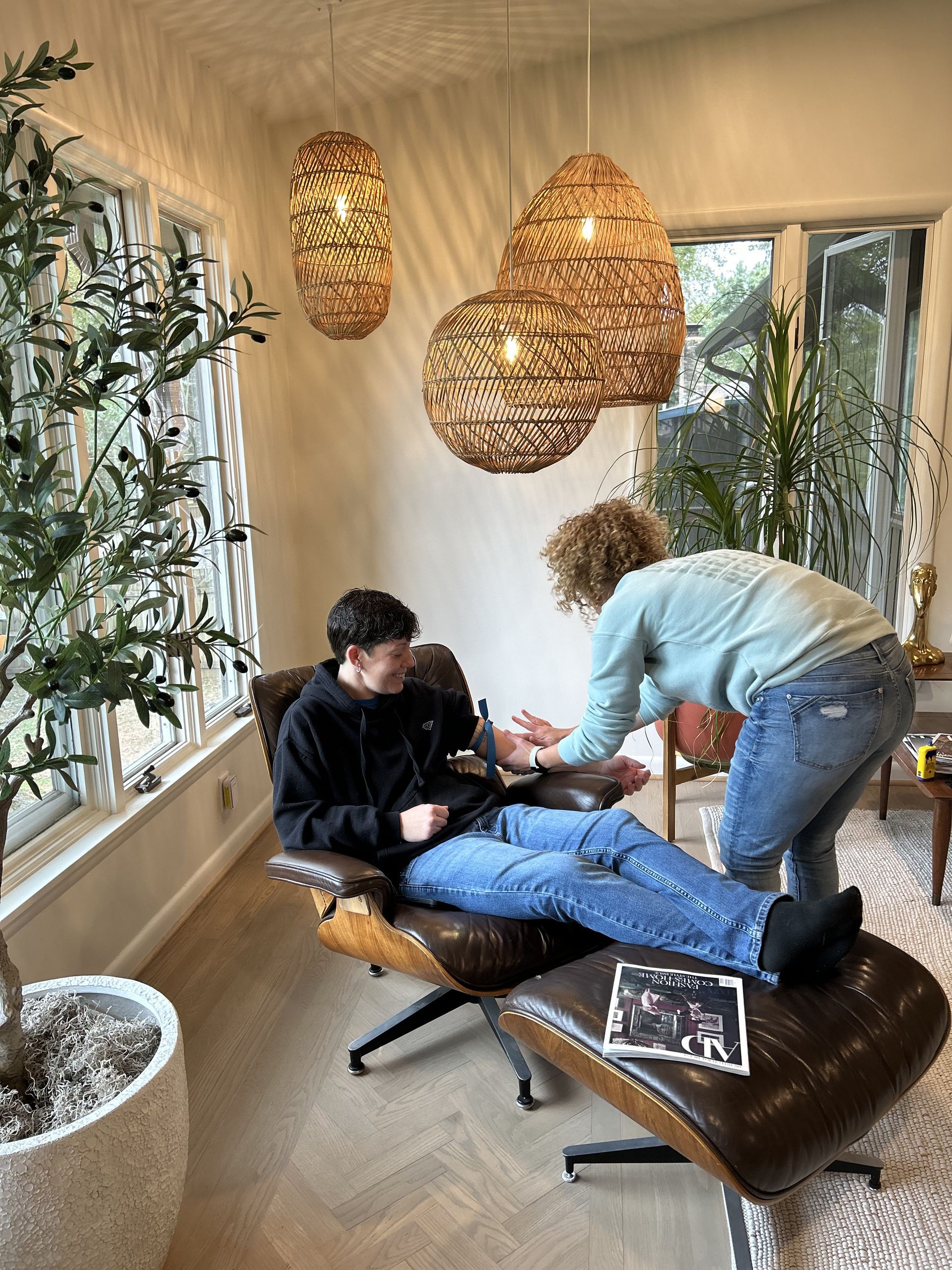 Person getting an IV in a bright room. A person assists, looking at arm. Modern lounge chair and hanging lights.