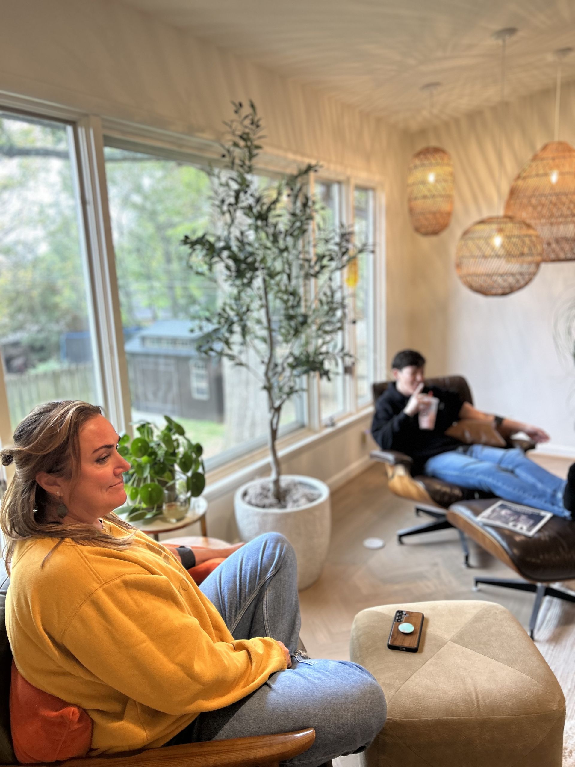 Woman and person lounge in bright living room, enjoying coffee. Large window, potted tree, and decorative lights.