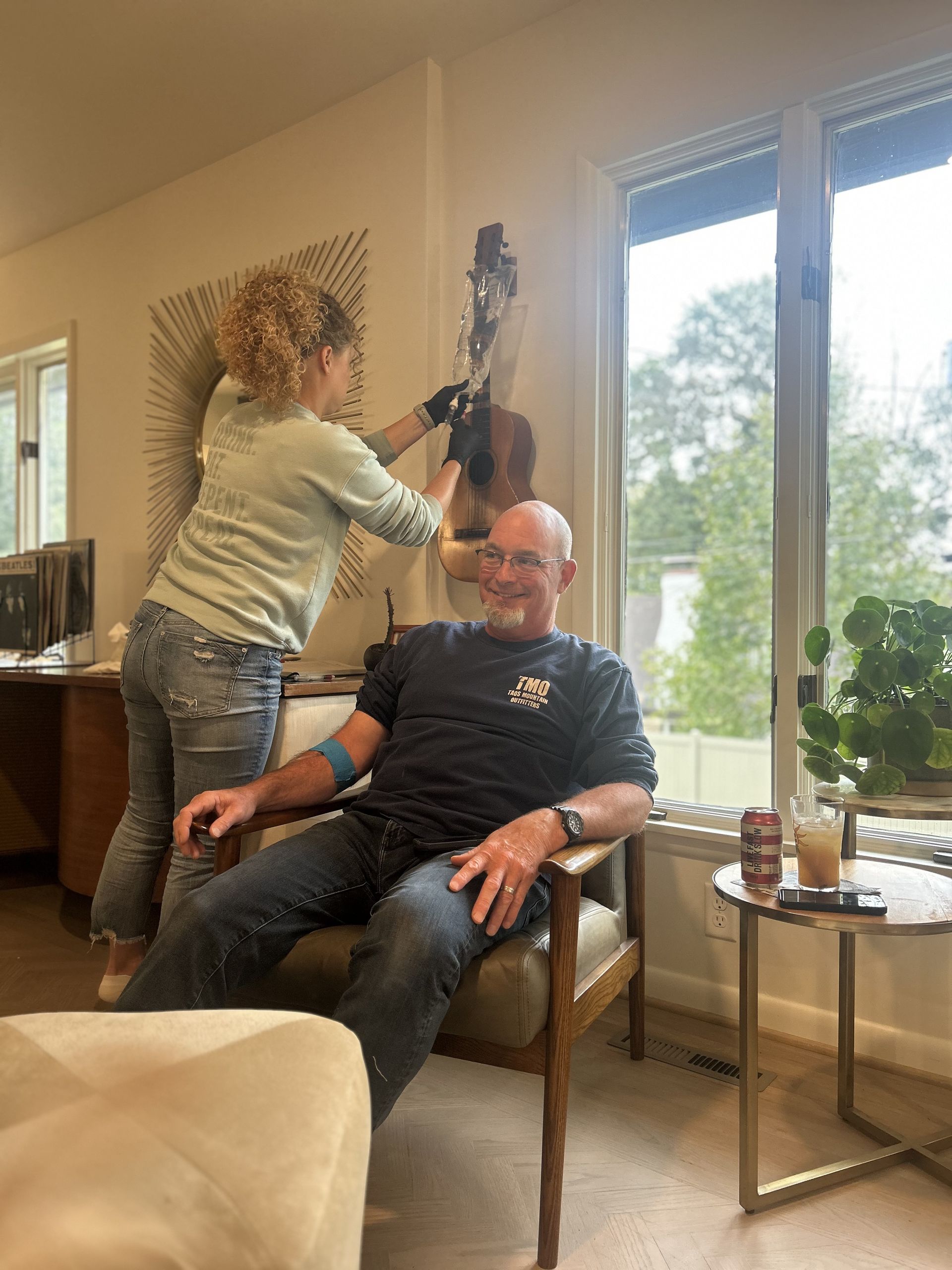 Woman adjusting instrument on wall, man seated in chair smiling, near window.