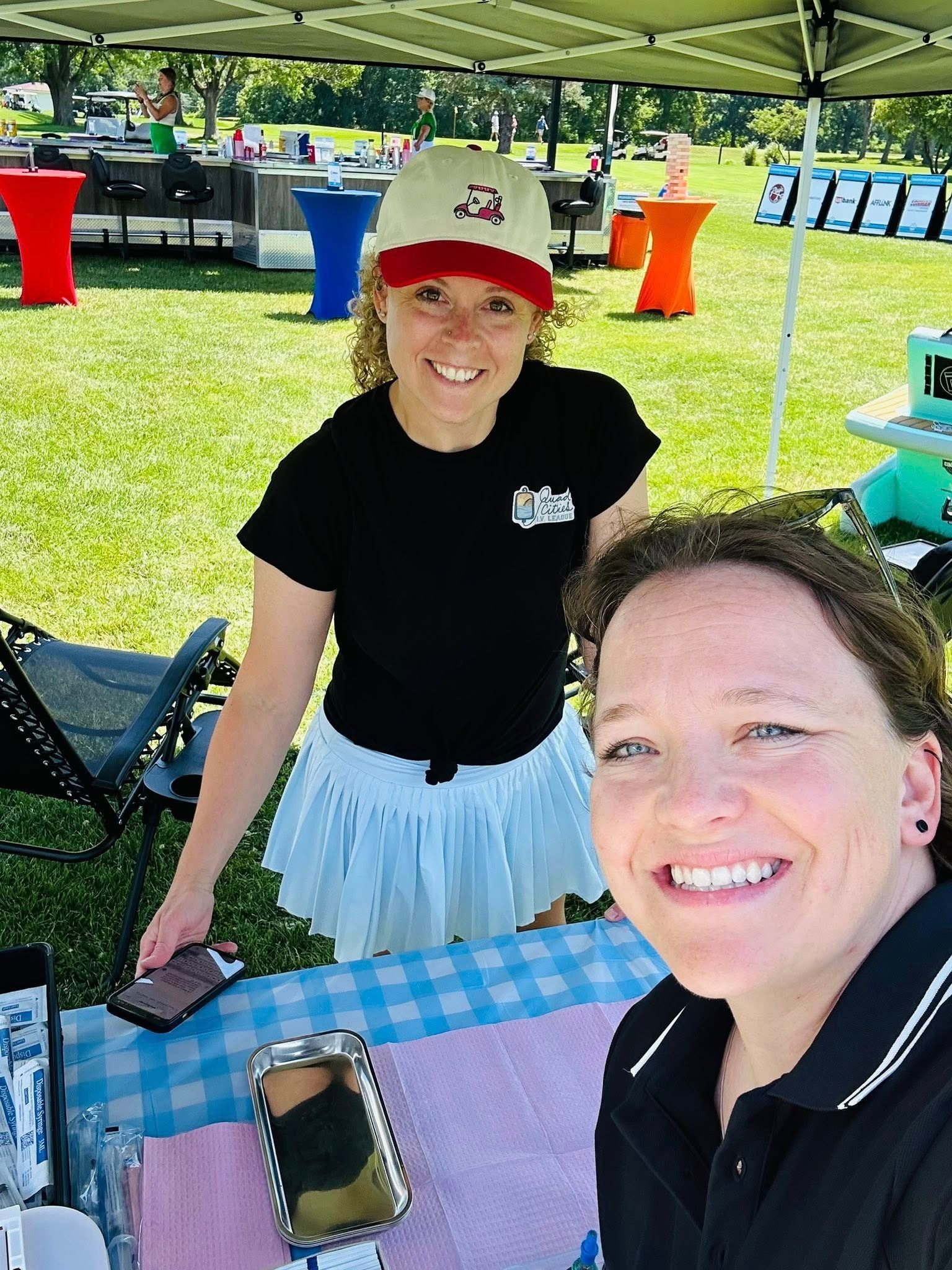 Two women smile at a table under a tent at a golf tournament. One wears a hat and skirt, the other takes a selfie. Outdoor setting.
