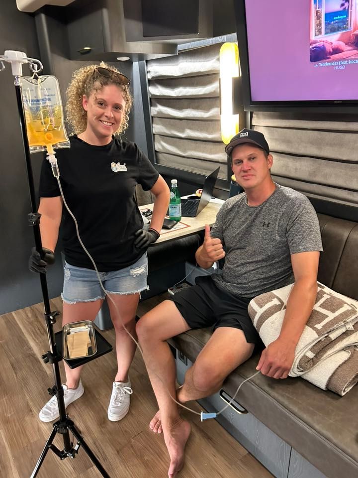A woman and Jon Pardi inside a trailer. Pardi receives IV fluids while giving a thumbs-up.