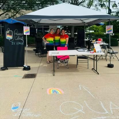 Three people in rainbow shirts stand at a pride event booth under a canopy. Chalk art, signs.