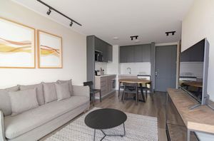 Modern apartment interior: grey sofa, artwork, kitchen with cabinets, dining table, and TV.