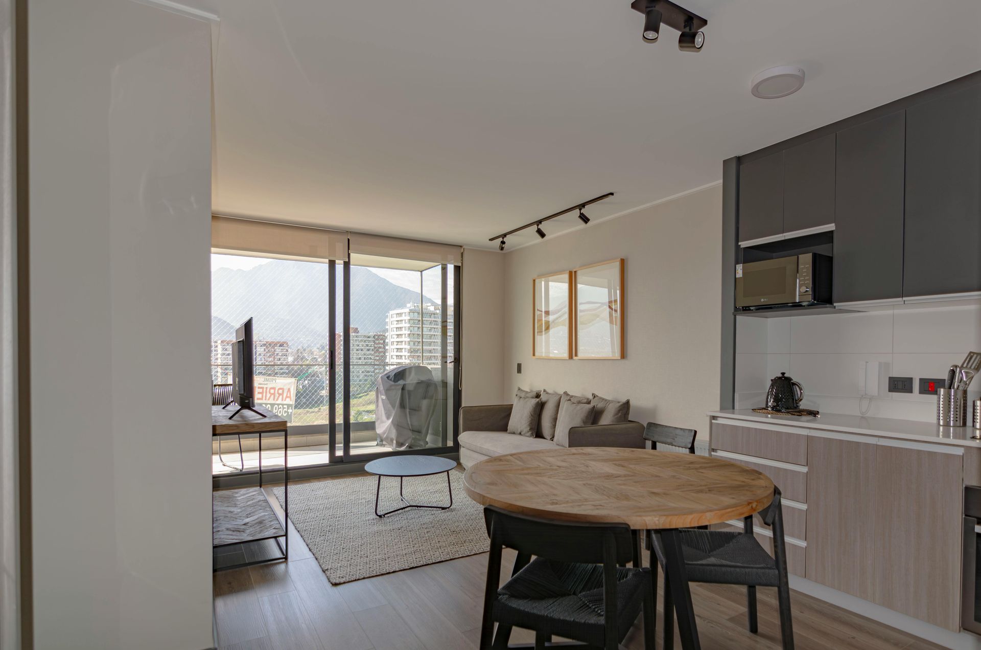 Modern apartment interior with a living area, kitchen, and dining table, with large windows overlooking a cityscape.