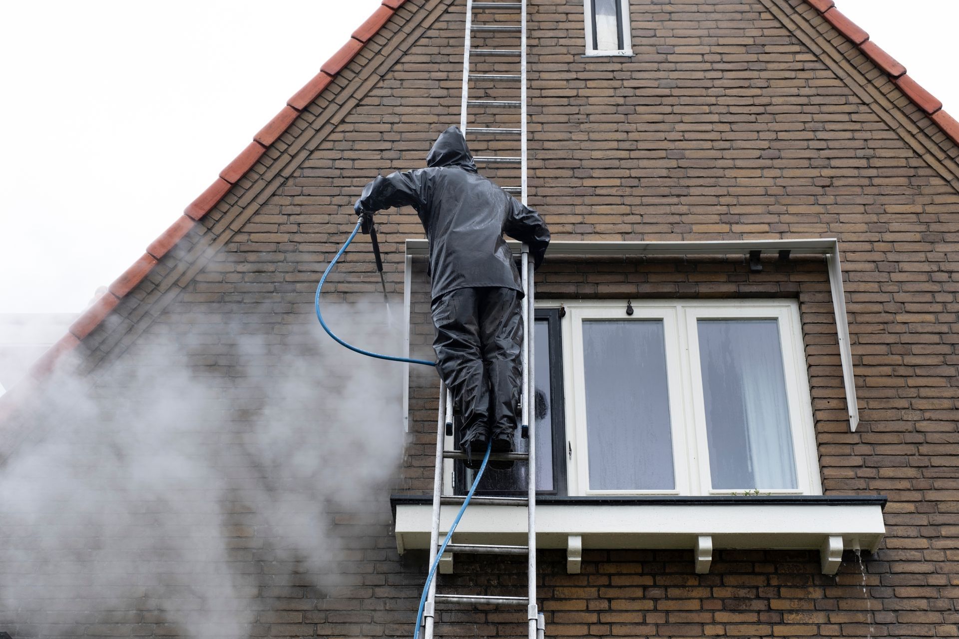 Person in waterproof suit on ladder, cleaning brick house with pressure washer, steam visible.
