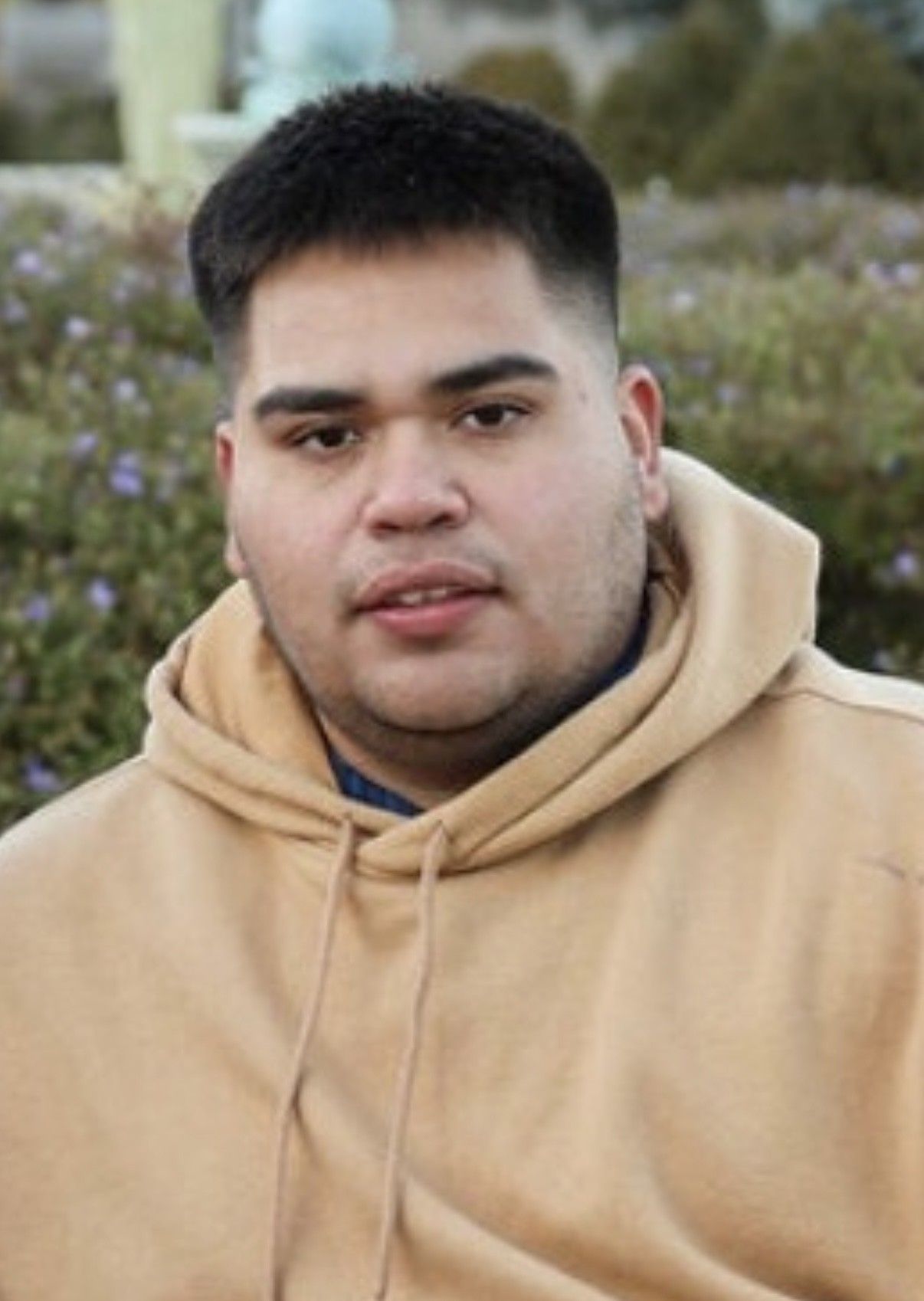 Man in a tan hoodie, short dark hair, looking at the camera. Outdoor setting, neutral expression.