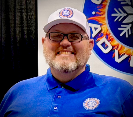 Man wearing glasses, hat, and blue shirt smiles in front of a logo.