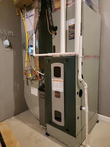 A furnace and attached humidifier in a gray room. White PVC pipes are visible, as well as gas lines.