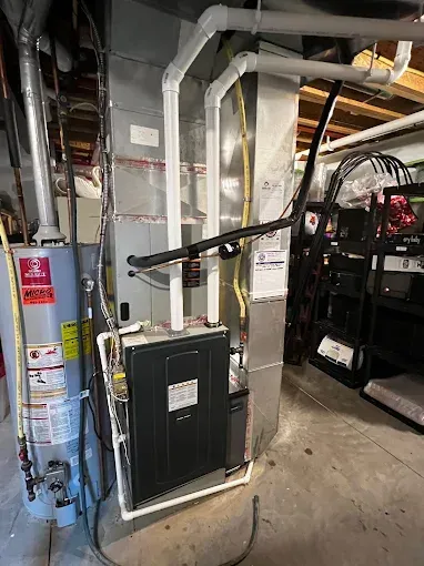 A furnace and water heater in a basement. The furnace is black, with white pipes. The water heater is silver.