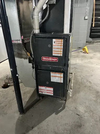 A Goodman furnace in a basement setting, with black exterior, ductwork, and warning labels.