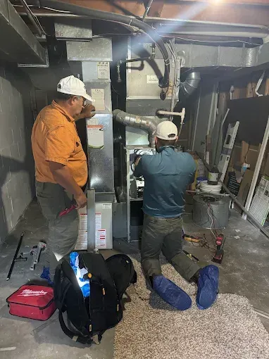 Two technicians working on HVAC unit in a basement. One kneels, the other observes.
