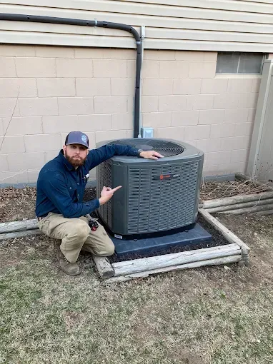 Man kneeling next to a central air conditioner, pointing at it, outdoors.