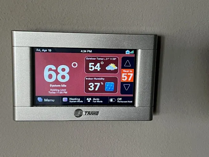 A silver Trane thermostat displaying 68 degrees, with an outdoor temperature of 54, humidity, and a set temperature of 57.