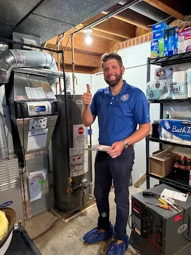 Man in blue shirt gives thumbs-up near a water heater and furnace. He is in a basement.