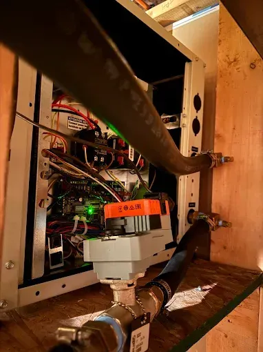 Interior of electrical equipment with wiring, pipes, and control valve against a wood wall.