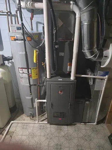 Furnace and water heater in a basement. Grey furnace, silver water heater, white pipes, tiled floor.