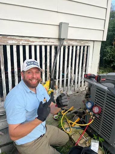 HVAC technician smiling while working on an air conditioner outside a building. He is wearing gloves and pointing.