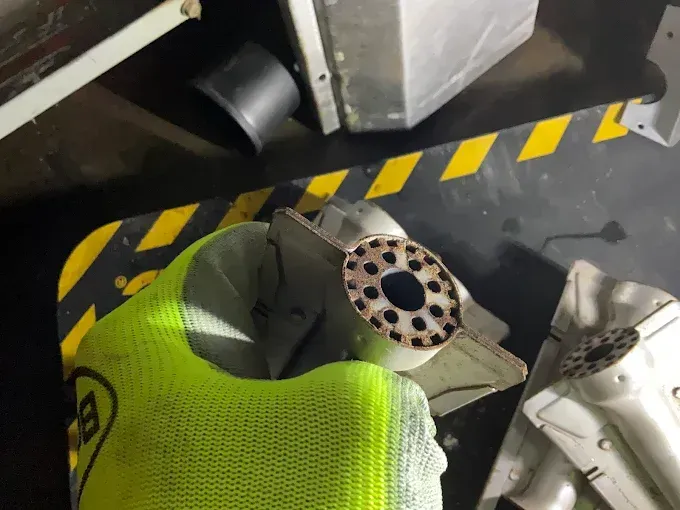 A gloved hand holds a metal component with multiple holes and a black center, with debris on a yellow and black surface.